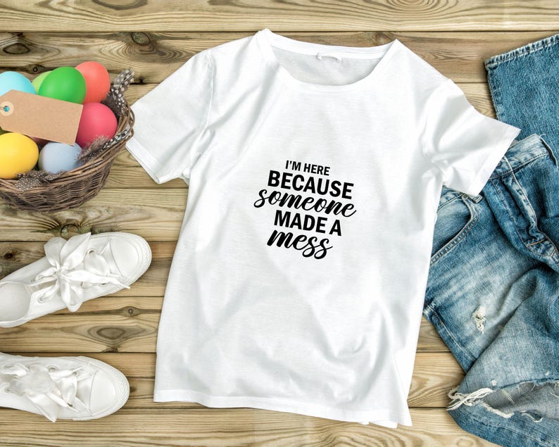May include: White t-shirt with black text that reads "I'm here because someone made a mess". The shirt is laid out on a wooden surface with a basket of colourful Easter eggs and a pair of blue jeans.