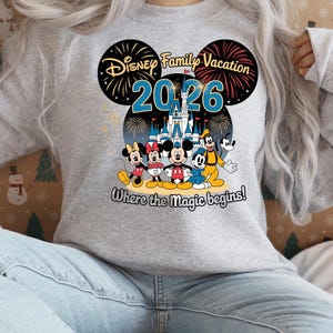 May include: A light gray sweatshirt featuring a colorful graphic of Disney characters, including Mickey and Minnie Mouse, Goofy, and Donald Duck. The design includes the text "Disney Family Vacation 2026" and "Where the Magic begins!"