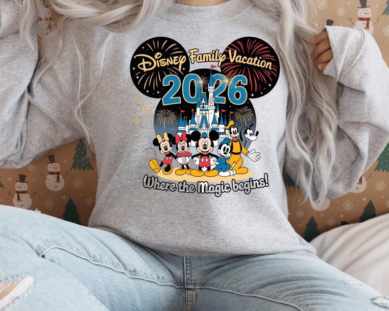 May include: A light gray sweatshirt featuring a colorful graphic of Disney characters, including Mickey and Minnie Mouse, Goofy, and Donald Duck. The design includes the text "Disney Family Vacation 2026" and "Where the Magic begins!"