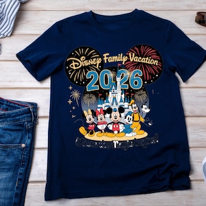 May include: Navy blue t-shirt featuring a Disney Family Vacation design for 2026. The graphic includes Mickey, Minnie, Donald, Goofy, and fireworks. The text "Where the magic begins!" is also included.
