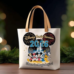 May include: Cream-colored tote bag with brown handles featuring a Disney Family Vacation design. The design includes Mickey Mouse, Minnie Mouse, Donald Duck, and Goofy, with the text "2026" and "Where the magic begins!"