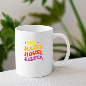May include: White ceramic mug with a colourful graphic that reads "The Happy House Keeper" in a retro font.