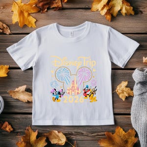 May include: White t-shirt with a Disney Trip design. The design includes the text "Disney Trip 2026" in gold, along with images of Mickey and Minnie Mouse, fireworks, and a castle. The shirt is displayed on a wooden surface with autumn leaves.