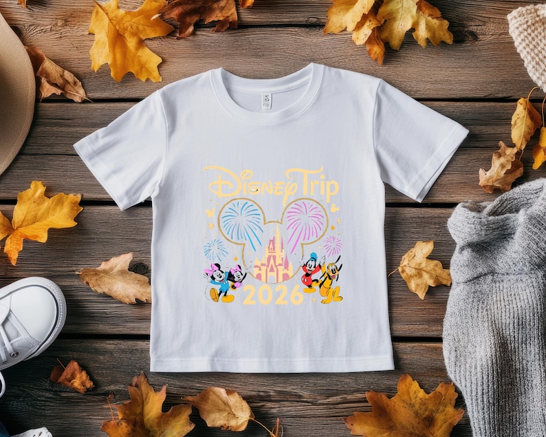 May include: White t-shirt with a Disney Trip design. The design includes the text "Disney Trip 2026" in gold, along with images of Mickey and Minnie Mouse, fireworks, and a castle. The shirt is displayed on a wooden surface with autumn leaves.