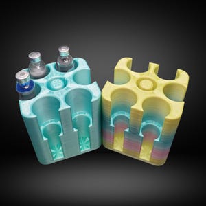 Puede incluir: Dos soportes para viales impresos en 3D. Uno es azul claro y contiene tres viales. El otro es amarillo con un degradado arcoíris y está vacío. Ambos soportes tienen seis aberturas circulares para viales.