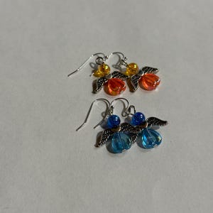 Pu&ograve; includere: Due paia di orecchini pendenti. Un paio presenta perline arancioni e gialle con ali d'angelo color argento. L'altro paio ha perline blu e ali d'angelo color argento. Entrambi i paia hanno ganci per le orecchie color argento.