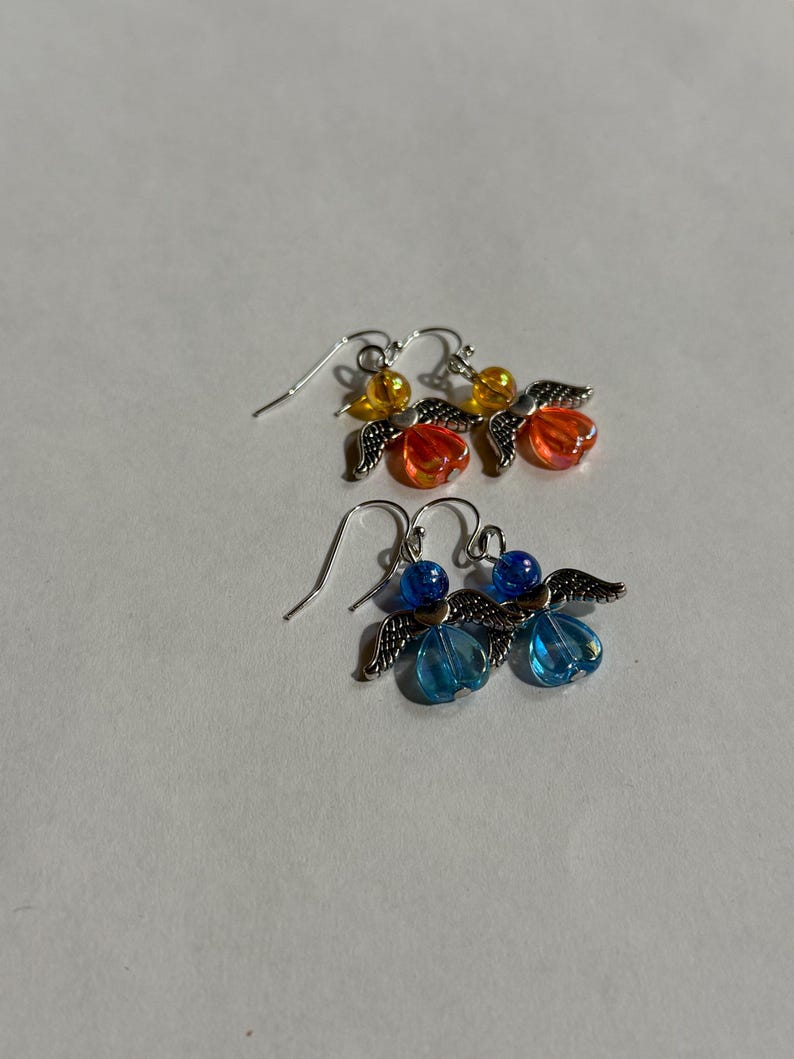 Pu&ograve; includere: Due paia di orecchini pendenti. Un paio presenta perline arancioni e gialle con ali d'angelo color argento. L'altro paio ha perline blu e ali d'angelo color argento. Entrambi i paia hanno ganci per le orecchie color argento.