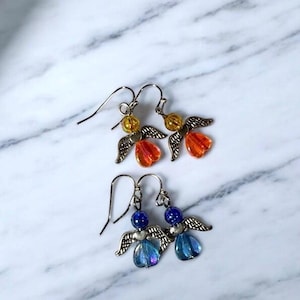 Pu&ograve; includere: Due paia di orecchini a forma di angelo fatti a mano. Un paio presenta perline arancioni e gialle con ali color argento. L'altro paio ha perline e ali blu. Gli orecchini sono esposti su una superficie di marmo bianco e grigio.