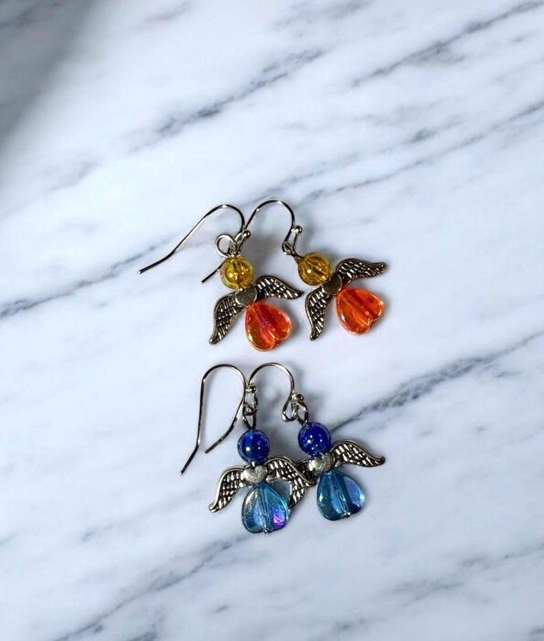Pu&ograve; includere: Due paia di orecchini a forma di angelo fatti a mano. Un paio presenta perline arancioni e gialle con ali color argento. L'altro paio ha perline e ali blu. Gli orecchini sono esposti su una superficie di marmo bianco e grigio.