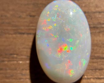 May include: An oval-shaped, white opal gemstone with iridescent flashes of red, green, and yellow. The polished stone displays a play of color, reflecting light in a spectrum of hues. The opal is set against a wooden surface.