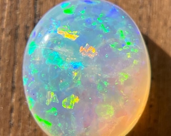 May include: A white opal gemstone with green, blue, and yellow flashes of color. The opal is oval shaped and has a smooth, polished surface.