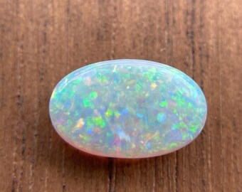 May include: An oval-shaped opal gemstone with a milky white base and iridescent flashes of green, blue, and yellow. The polished stone is set against a brown wooden surface, showcasing its natural beauty and color variations. This gemstone is suitable for jewelry making.