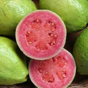 May include: Close-up of several green guavas, some whole and others sliced open to reveal a vibrant pink interior with small seeds. The fruits are arranged in a woven basket, showcasing their natural colors and textures.