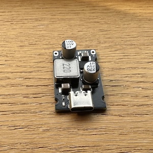 Puede incluir: Una pequeña placa de circuito electrónica negra con varios componentes, incluyendo condensadores marcados con "220" y "100 35V UT". Cuenta con un puerto USB-C y está sobre una superficie de madera.