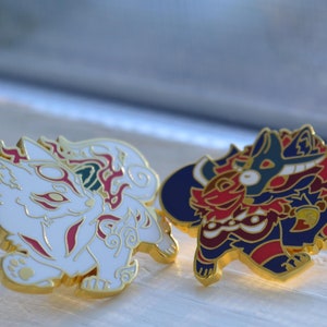 May include: Two enamel pins featuring stylized fox and canine designs. The fox pin is white with red and gold accents. The canine pin is red, blue, and gold. Both pins have gold borders and are set against a light background.