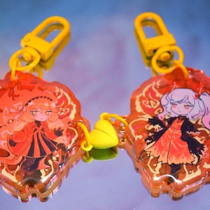 Könnte beinhalten: Zwei Schlüsselanhänger im Anime-Stil mit gelben Verschlüssen. Jeder Schlüsselanhänger zeigt einen Cartoon-Charakter in einem roten und orangefarbenen Design. Die Schlüsselanhänger sind durch einen gelben herzförmigen Anhänger verbunden.