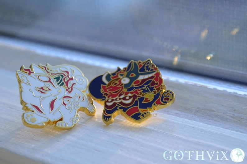 May include: Two enamel pins featuring stylized fox and canine designs. The fox pin is white with red and gold accents. The canine pin is red, blue, and gold. Both pins have gold borders and are set against a light background.