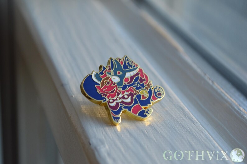 May include: A colorful enamel pin featuring a stylized creature with a blue and red color scheme, accented with gold. The pin has a detailed design and is set against a light background. The pin is a collectible item.