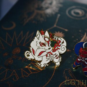 May include: A white and red fox enamel pin with a gold border, featuring a stylized design. The pin is set against a dark teal background with gold celestial patterns. A second, colorful pin is visible to the right.