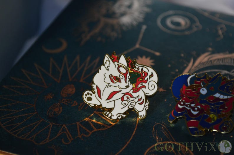 May include: A white and red fox enamel pin with a gold border, featuring a stylized design. The pin is set against a dark teal background with gold celestial patterns. A second, colorful pin is visible to the right.