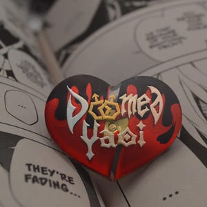 K&ouml;nnte beinhalten: Eine rot-schwarze herzf&ouml;rmige Anstecknadel mit den Worten "Doomed Yaoi" in wei&szlig;er und goldener Schrift. Die Nadel ist in zwei H&auml;lften gebrochen, wobei die beiden H&auml;lften leicht versetzt sind.