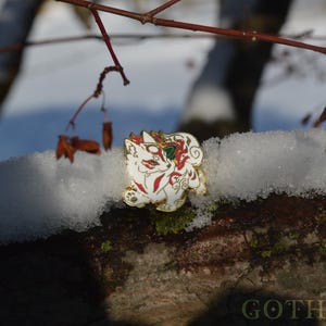 May include: A white and gold enamel pin featuring a stylized fox design with red and green accents. The pin is set against a snowy, natural background with tree branches.