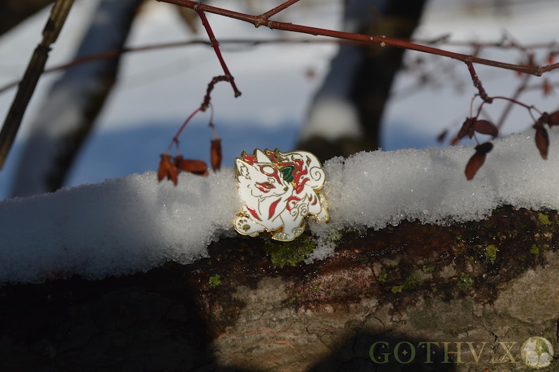 May include: A white and gold enamel pin featuring a stylized fox design with red and green accents. The pin is set against a snowy, natural background with tree branches.