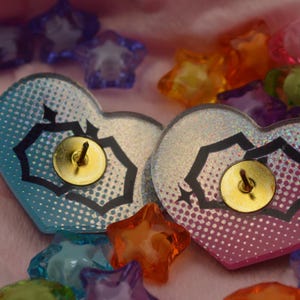 K&ouml;nnte beinhalten: Zwei regenbogenfarbene herzf&ouml;rmige Pins mit schwarzen geometrischen Mustern und goldenen Stecknadeln. Die Pins befinden sich auf einer rosa Oberfl&auml;che mit bunten sternf&ouml;rmigen Verzierungen.