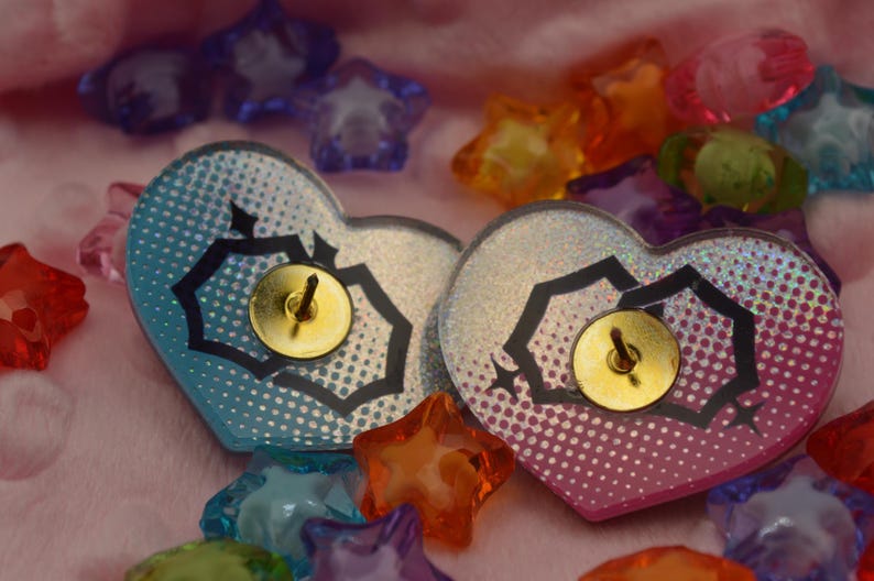 K&ouml;nnte beinhalten: Zwei regenbogenfarbene herzf&ouml;rmige Pins mit schwarzen geometrischen Mustern und goldenen Stecknadeln. Die Pins befinden sich auf einer rosa Oberfl&auml;che mit bunten sternf&ouml;rmigen Verzierungen.