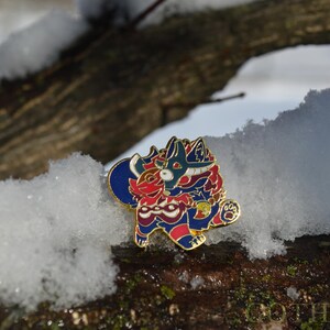 May include: A colorful enamel pin featuring a stylized creature design in red, blue, and gold. The pin is set against a snowy, natural background, highlighting its intricate details and vibrant colors. The pin is a collectible item.