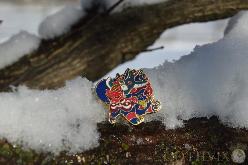 May include: A colorful enamel pin featuring a stylized creature design in red, blue, and gold. The pin is set against a snowy, natural background, highlighting its intricate details and vibrant colors. The pin is a collectible item.