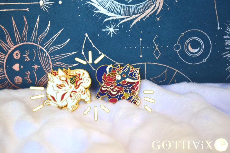 May include: Two enamel pins featuring stylized fox designs. One is white with red and gold accents, the other is red, blue, and gold. The pins are on a fluffy white surface with a dark teal background with celestial designs.