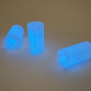 May include: Three glowing blue cylindrical objects are arranged on a light-colored surface. The objects appear to be made of a translucent material, emitting a bright, cool blue light. The cylinders are of varying orientations.