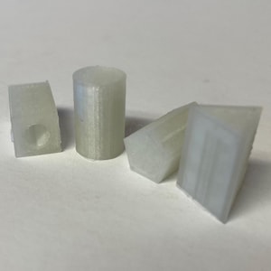 May include: A collection of four off-white, 3D-printed plastic objects. One is cylindrical, and the others are angular, with one featuring a hole. The objects are arranged on a plain, light-colored surface.