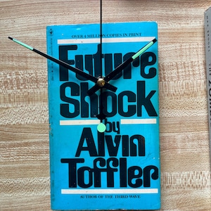May include: A clock made from a book titled "Future Shock" by Alvin Toffler. The book cover is light blue with black text and clock hands. The clock hands are black with green tips and a gold center.