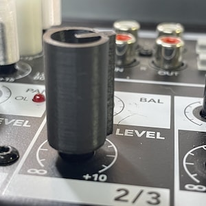 May include: Close-up of a black audio mixer with various knobs and inputs. The mixer features a large, cylindrical control knob labeled "LEVEL" and a red indicator light. The panel has white text and markings.