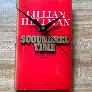 May include: A rectangular clock made from a book cover. The cover is bright red with the title "SCOUNDREL TIME" in gold lettering. The author's name, Lillian Hellman, is in white. Black clock hands indicate the time.