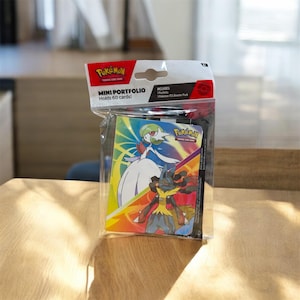 Puede incluir: Un paquete de plástico transparente que contiene un Mini Portfolio Pokémon. La cartera negra presenta imágenes coloridas de personajes Pokémon. El embalaje incluye el texto "MINI PORTFOLIO Holds 60 cards!" y "Pokémon Trading Card Game".