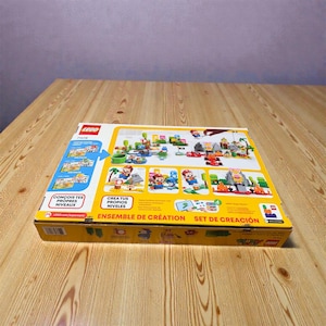 May include: A yellow LEGO Super Mario set box, featuring images of Mario characters and level designs. The box has text in multiple languages, including "CON&Ccedil;OIS TES PROPRES NIVEAUX" and "ENSEMBLE DE CR&Eacute;ATION". The box is on a wooden surface.