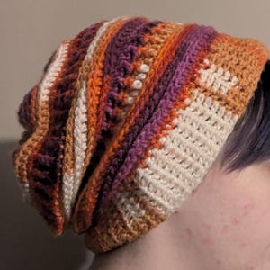 Op de afbeelding: Een gebreide beanie met strepen in wit, oranje, bruin en paars.