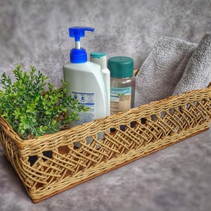 Puede incluir: Una cesta de mimbre tejida con un acabado marrón claro contiene una botella de jabón de manos azul y blanco, una botella transparente con tapa verde y dos toallas grises. La cesta está sobre una superficie gris.