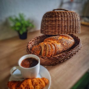 Op de afbeelding: Een witte keramische koffiekop met een bruine vloeistof erin, een croissant en een rieten broodmand met gesneden brood. De broodmand heeft een deksel.