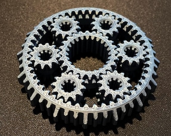 Op de afbeelding: Een set van in elkaar grijpende tandwielen gemaakt van grijs plastic. De tandwielen zijn in een cirkel gerangschikt, met het grootste tandwiel in het midden. De tandwielen zijn allemaal van verschillende grootte en hebben een verschillend aantal tanden.