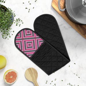Puede incluir: Un par de guantes de horno negros con un patrón geométrico rosa y gris en un lado. Los guantes están acolchados y tienen un forro resistente al calor.