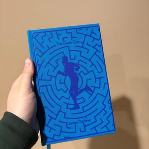 May include: A blue book with a maze pattern and a silhouette of a person running. The title "The Death Cure" and the author "James Dashner" are printed on the cover.