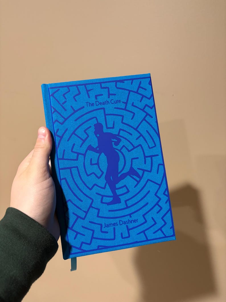 May include: A blue book with a maze pattern and a silhouette of a person running. The title "The Death Cure" and the author "James Dashner" are printed on the cover.