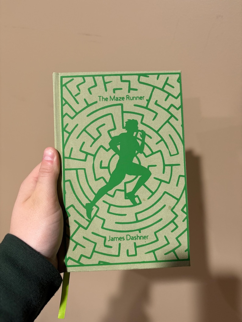 May include: A green paperback book with a maze pattern on the cover. The title "The Maze Runner" and the author "James Dashner" are printed on the cover.