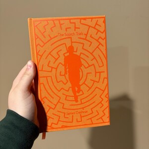 May include: An orange paperback book with the title "The Scorch Trials" and the author "James Dashner" printed on the cover. The cover features a silhouette of a person running through a maze.