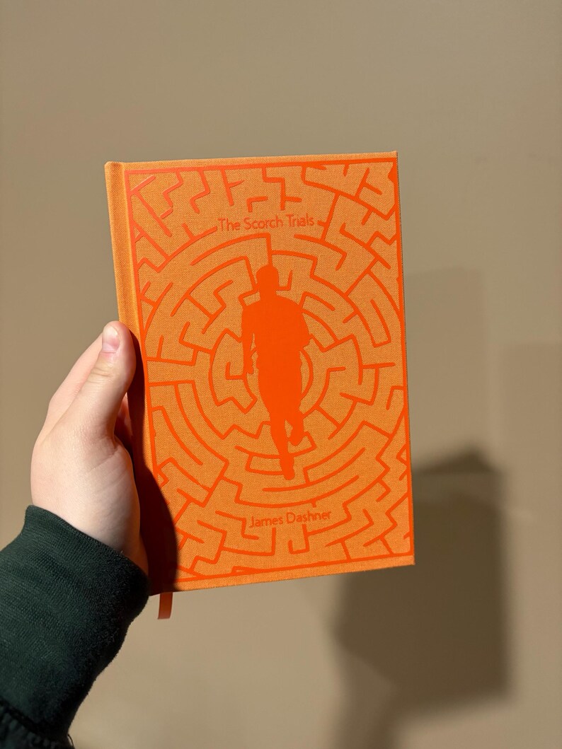 May include: An orange paperback book with the title "The Scorch Trials" and the author "James Dashner" printed on the cover. The cover features a silhouette of a person running through a maze.