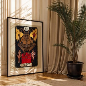 Puede incluir: Obra de arte enmarcada de una carta del tarot que representa al Diablo. La obra tiene un fondo negro con detalles dorados y rojos, y el texto "THE DEVIL" en la parte inferior. El marco es negro.
